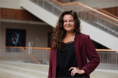 [ai] A woman with curly hair wearing a burgundy blazer and a black top, standing confidently in a modern building with a staircase in the background.